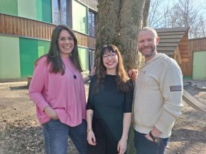 3 Menschen lächeln in die Kamera, hinter ihnen ein Baum und eine grün vertäfelte Kita