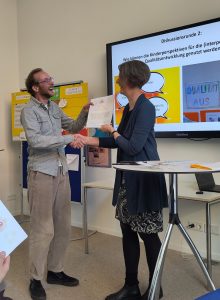 Eine Frau und ein Mann schütteln Hände, der Mann lacht und bekommt eine Urkunde übergeben.