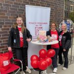 Drei Frauen lachen in die Kamera. Sie stehen an einem Bistro-Tisch an einer Backstein-Wand, davor rote Ballons und dahinter ein weißes Banner mit dem Logo der Pestalozzi-Stiftung Hamburg