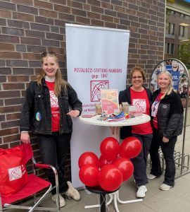 Drei Frauen lachen in die Kamera. Sie stehen an einem Bistro-Tisch an einer Backstein-Wand, davor rote Ballons und dahinter ein weißes Banner mit dem Logo der Pestalozzi-Stiftung Hamburg. Man erkennt am Fahrradständer, dass es bei einer Budni-Filiale ist.